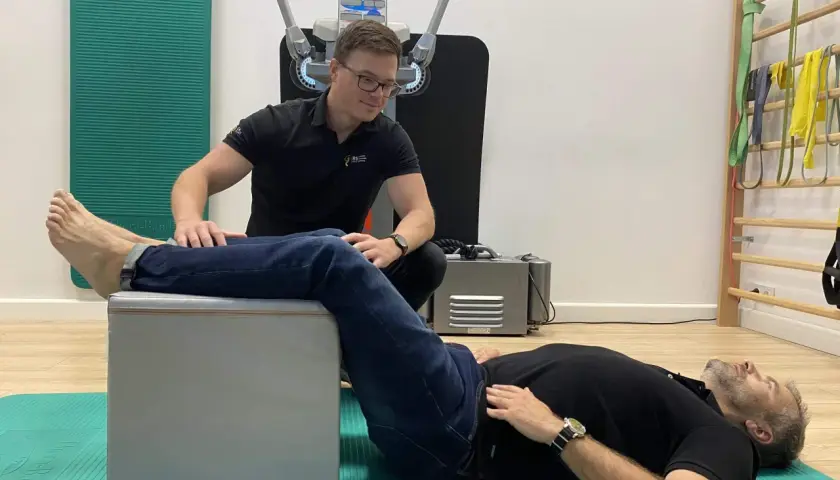 Fizjoterapeuta pomaga pacjentowi w ćwiczeniach na rwę kulszową. Pacjent leży na macie, z nogą opartą na podwyższeniu, a terapeuta wykonuje palpacje.